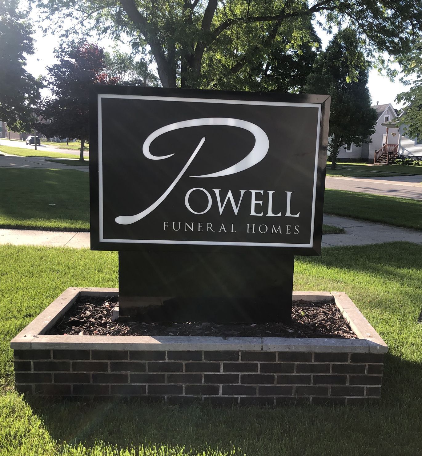 Sign for Powell Funeral Homes; black with white logo and text, set in a brick-lined bed of mulch.