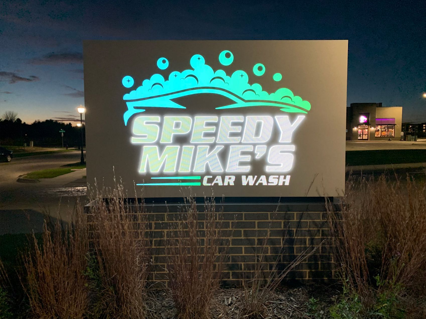 Sign for 'Speedy Mike's Car Wash' with illuminated bubbles and car graphic. Set against a dusk sky, near a brick wall.