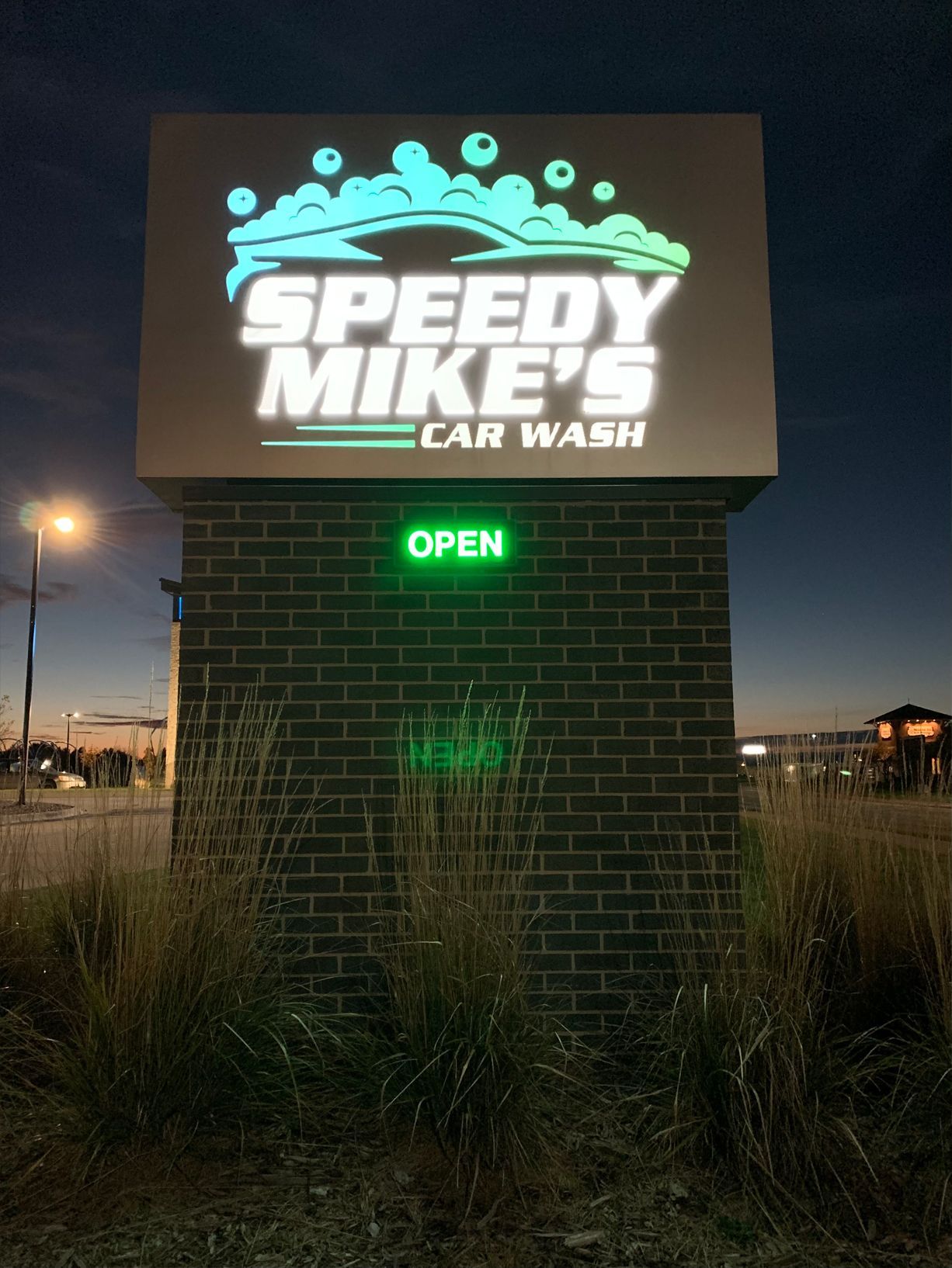 Speedy Mike's Car Wash sign at dusk, featuring illuminated logo, 'OPEN' sign, and brick pillar.