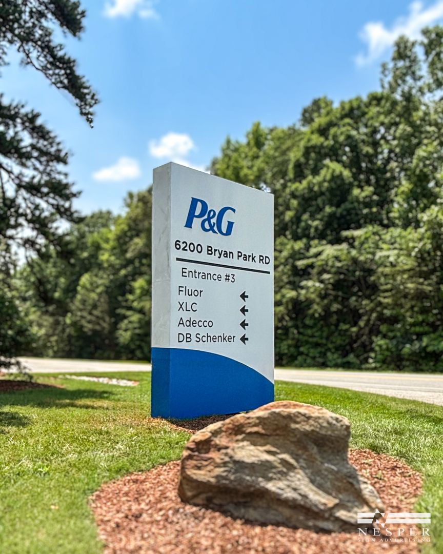 Sign for P&G with address and building directions, set in front of a green wooded area.