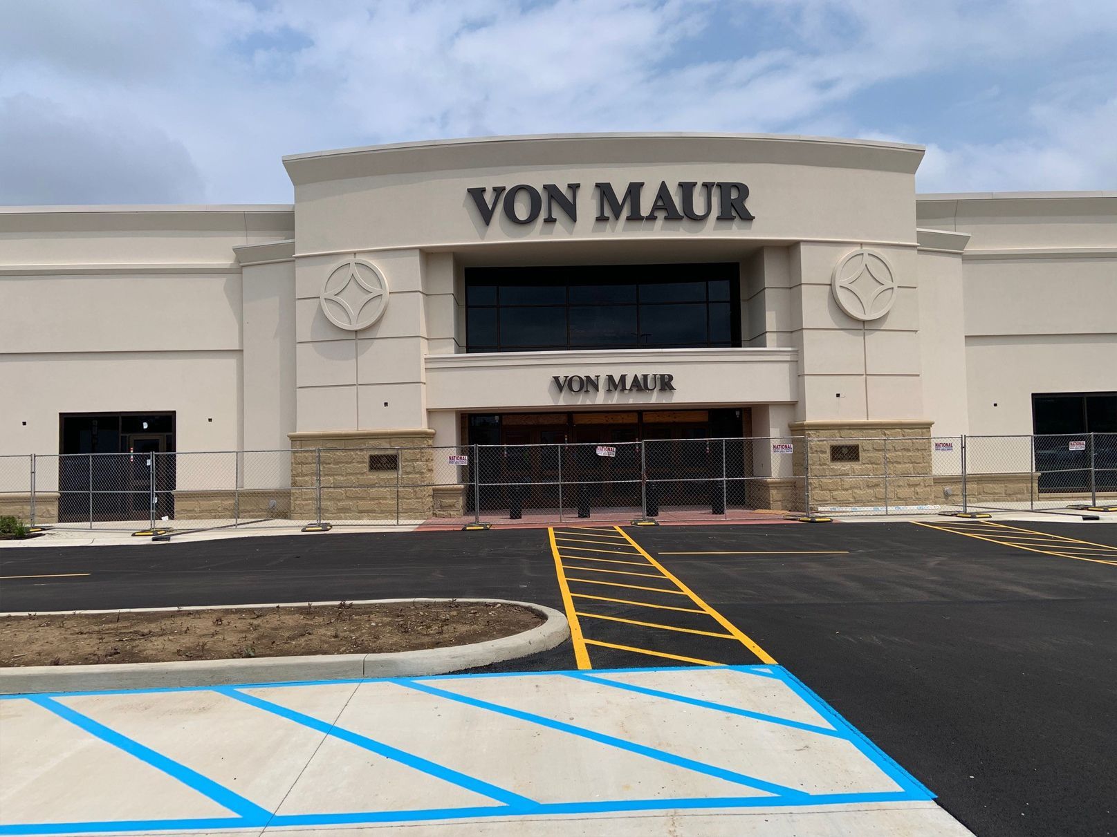 Von Maur department store exterior; tan building with black sign, construction fencing at entrance, blue curb paint.