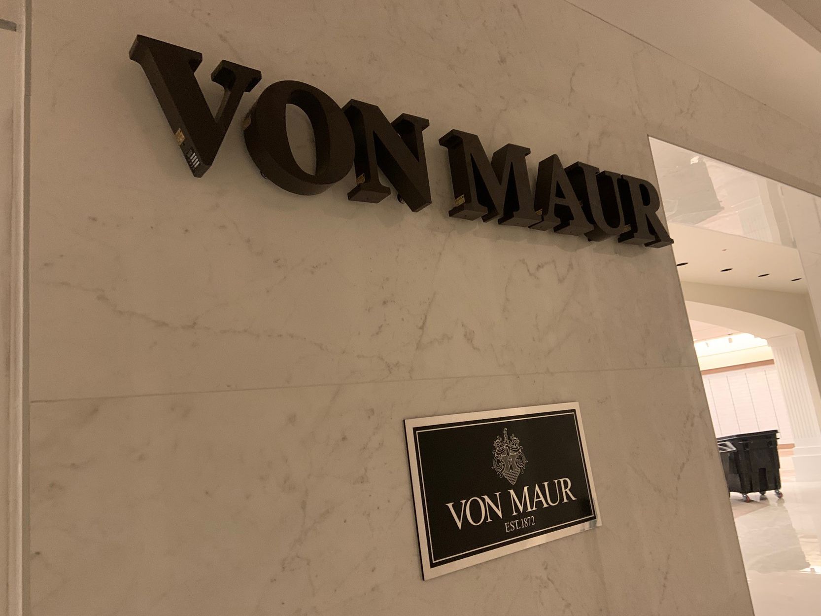 'Von Maur' store signage on a textured white wall; black letters and logo.