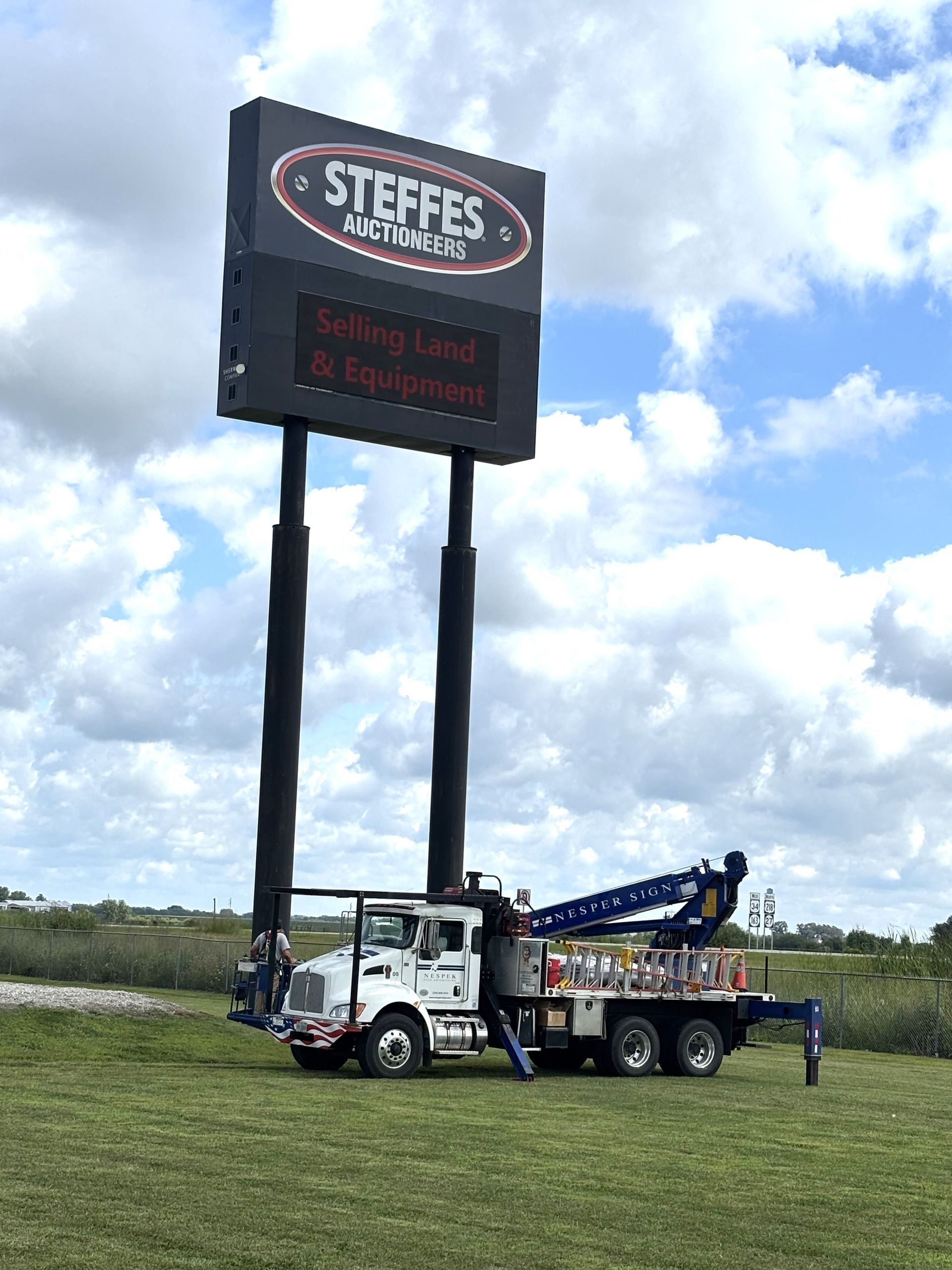 A white truck with a crane next to a tall sign for Steffes Electric; grassy field, cloudy sky.