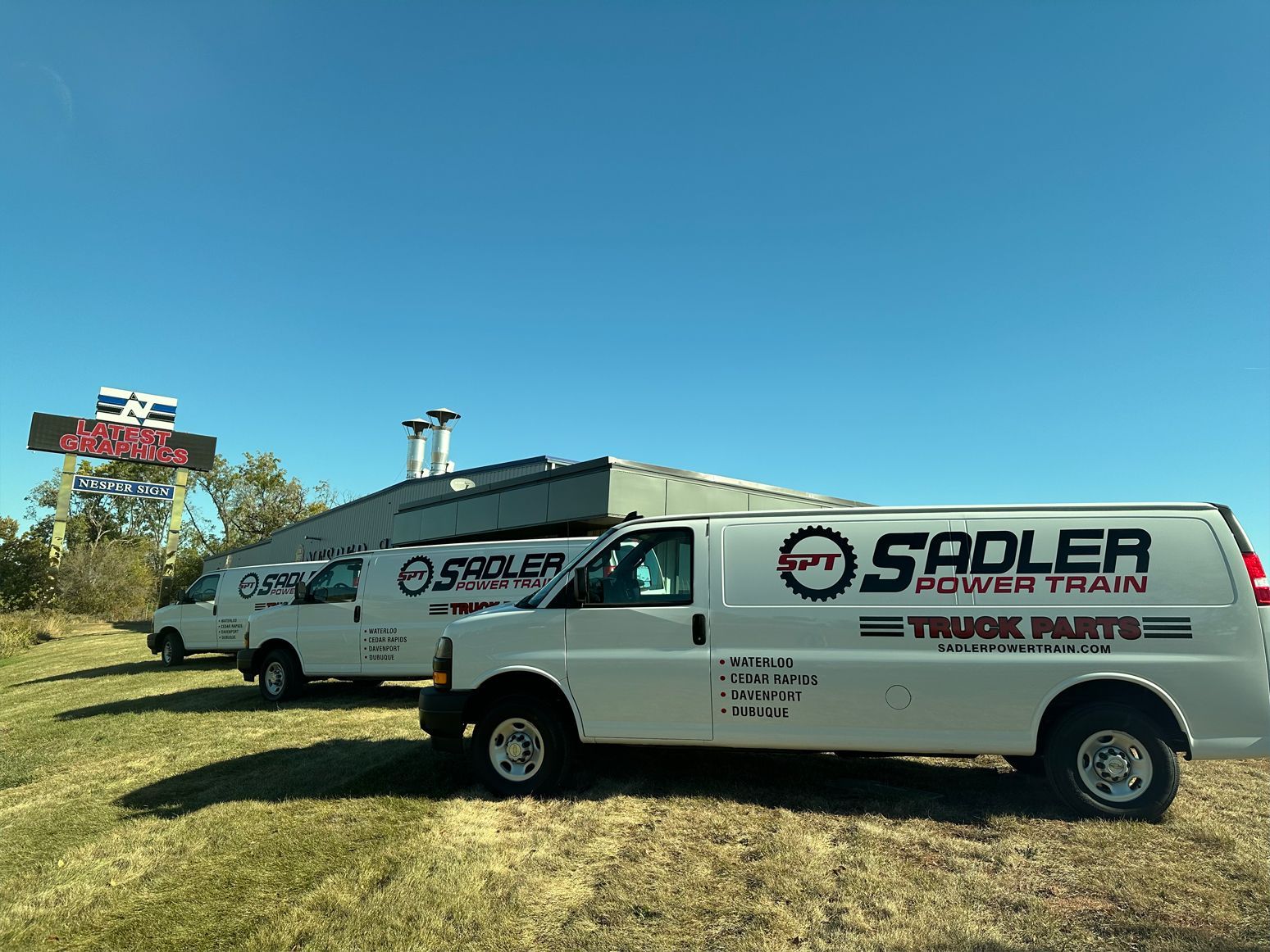 Three white work vans with 'Sadler Truck Parts' signage parked outside a building with a sign on a clear, blue day.