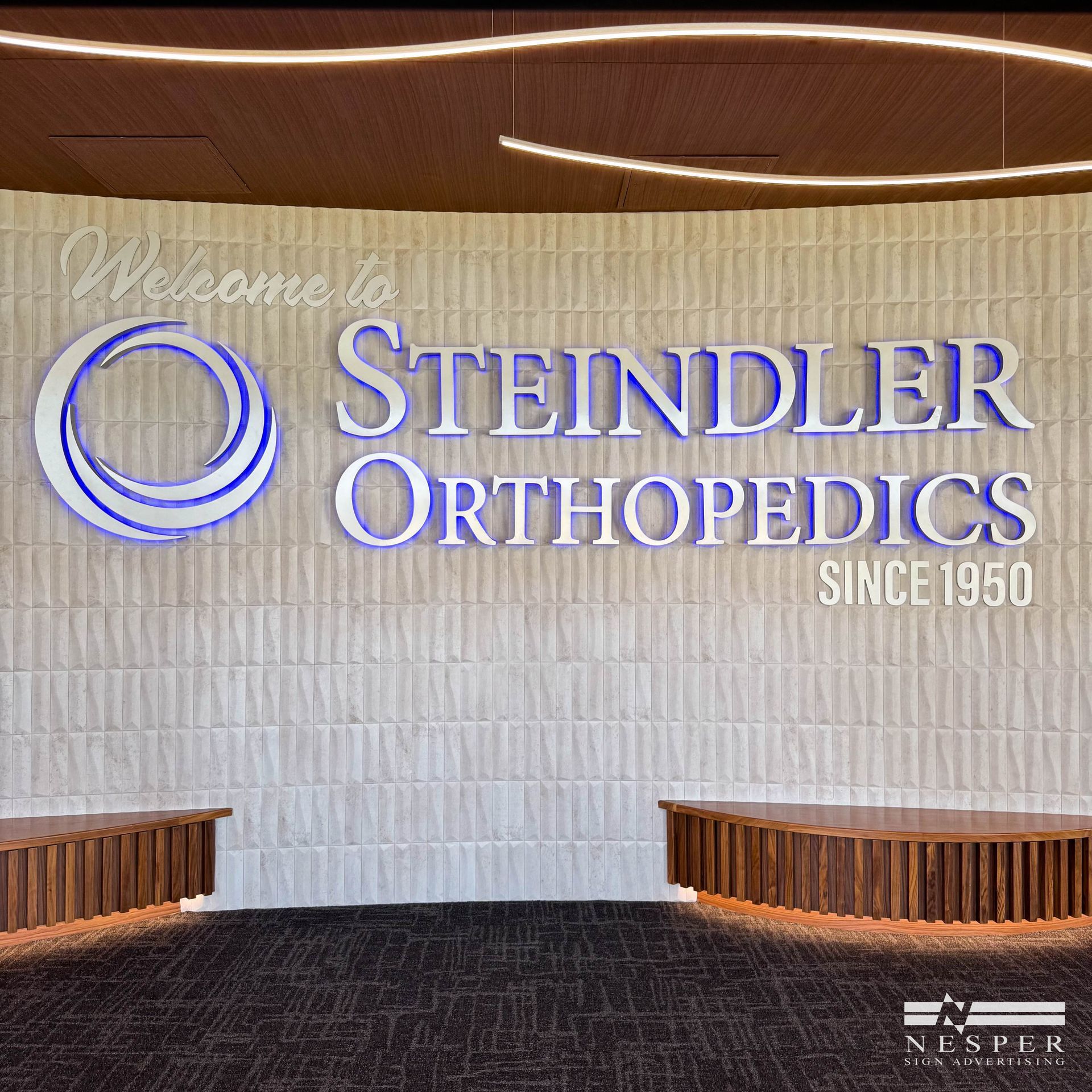 Welcome sign for Steindler Orthopedics, white text and blue logo on textured wall, with brown benches below.