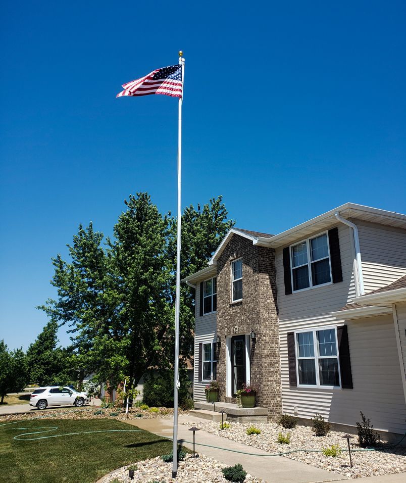 American flag waving on a tall pole in front of a two-story house with a blue sky.