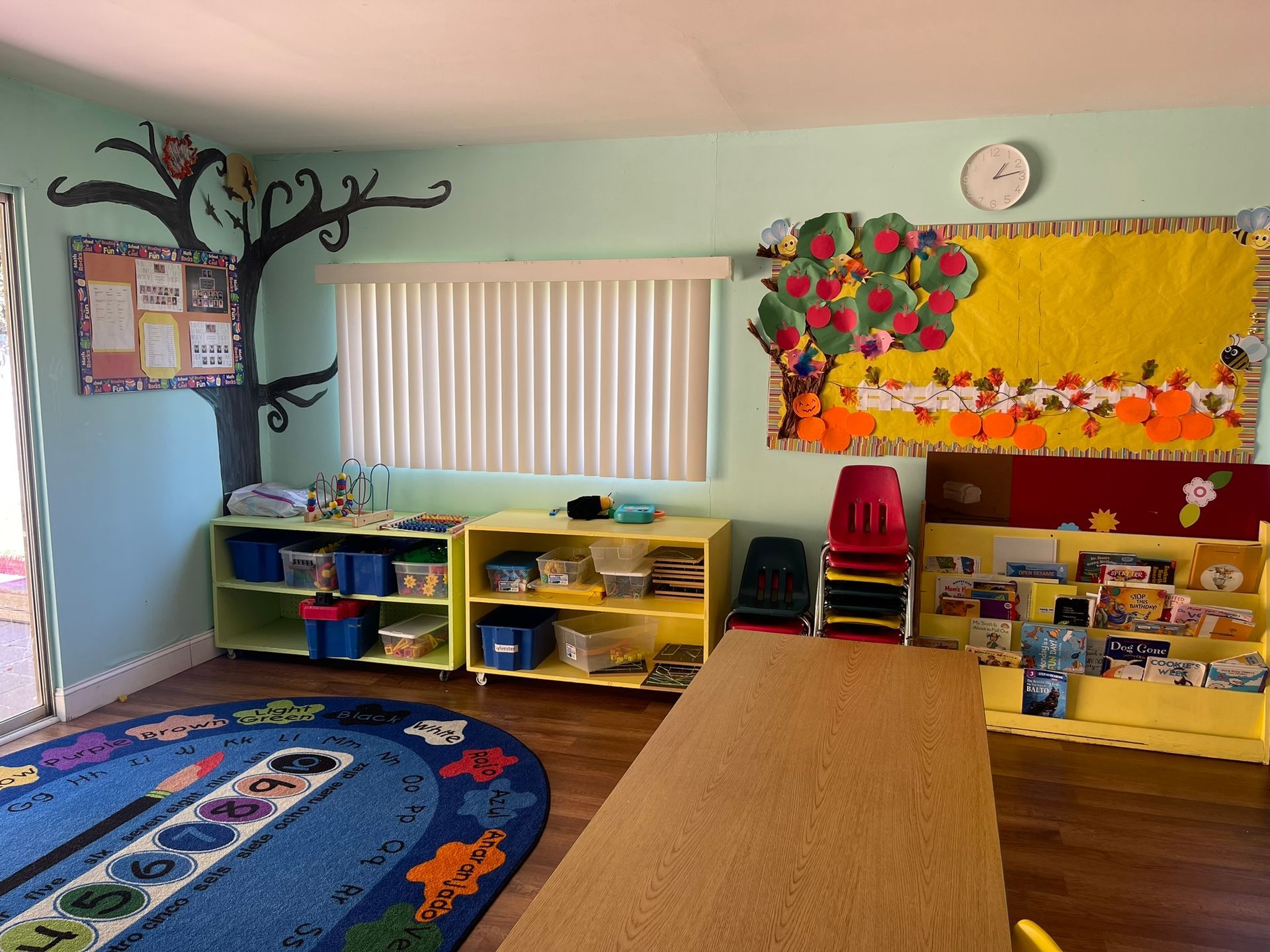 A classroom with a table , chairs , shelves and a clock on the wall.