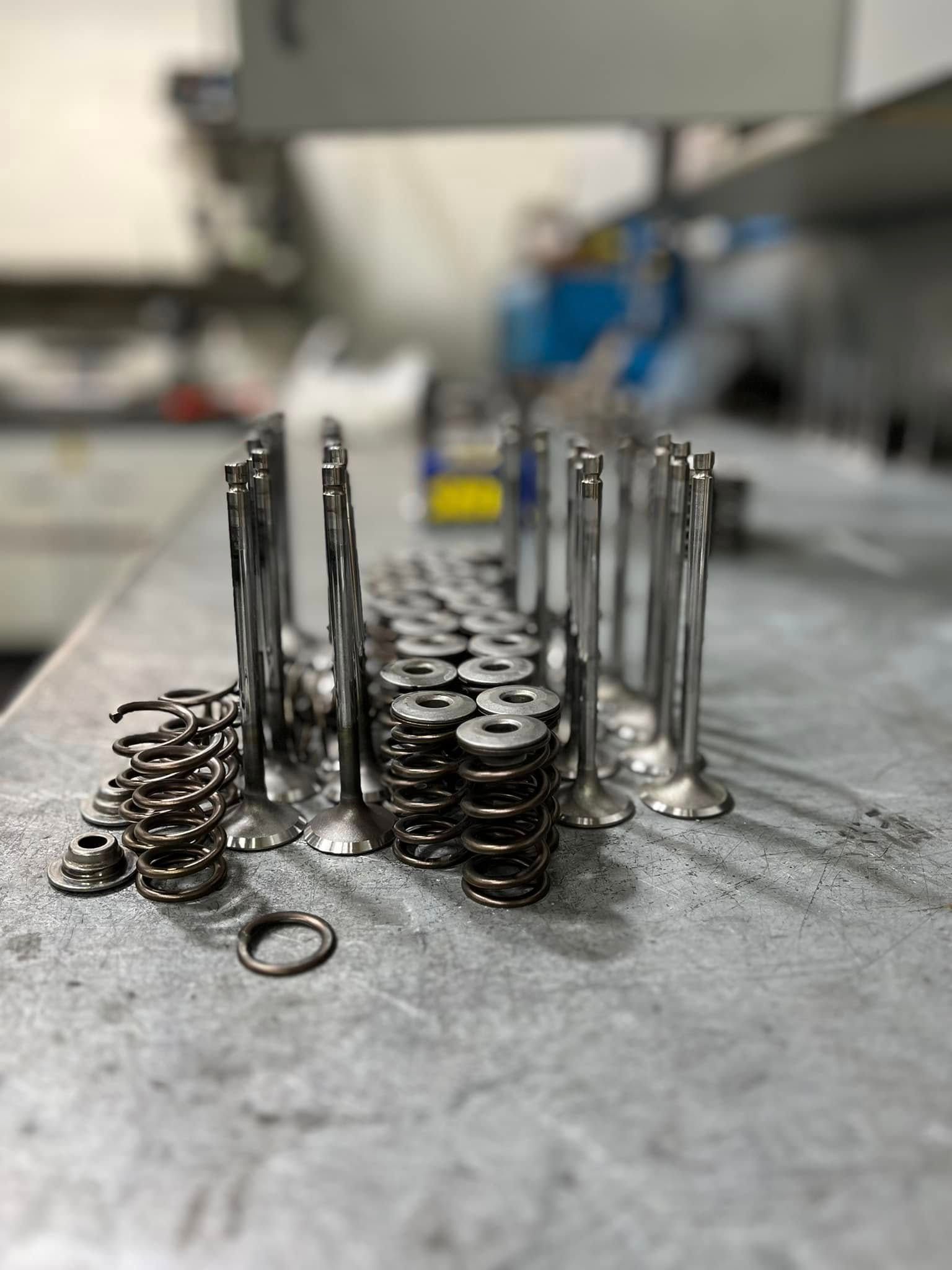 Several engine valves and coiled valve springs stand upright on a metal workbench in a workshop.