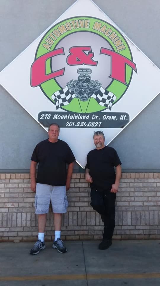 Two people stand in front of a diamond-shaped C&T Automotive Machine sign on a brick wall.
