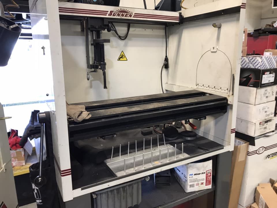 A white Sunnen industrial honing machine sits in a workshop, featuring a metal guide rack and a vertical operating handle.