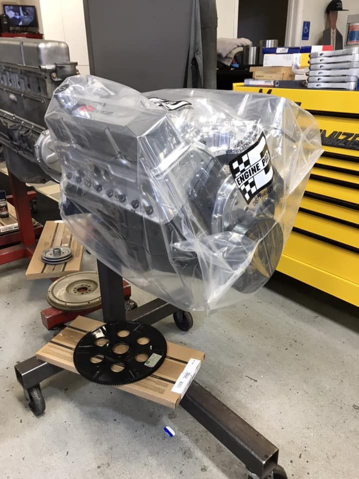 A new V8 engine on a mobile metal stand, wrapped in clear plastic, inside an automotive workshop with yellow tool chests.