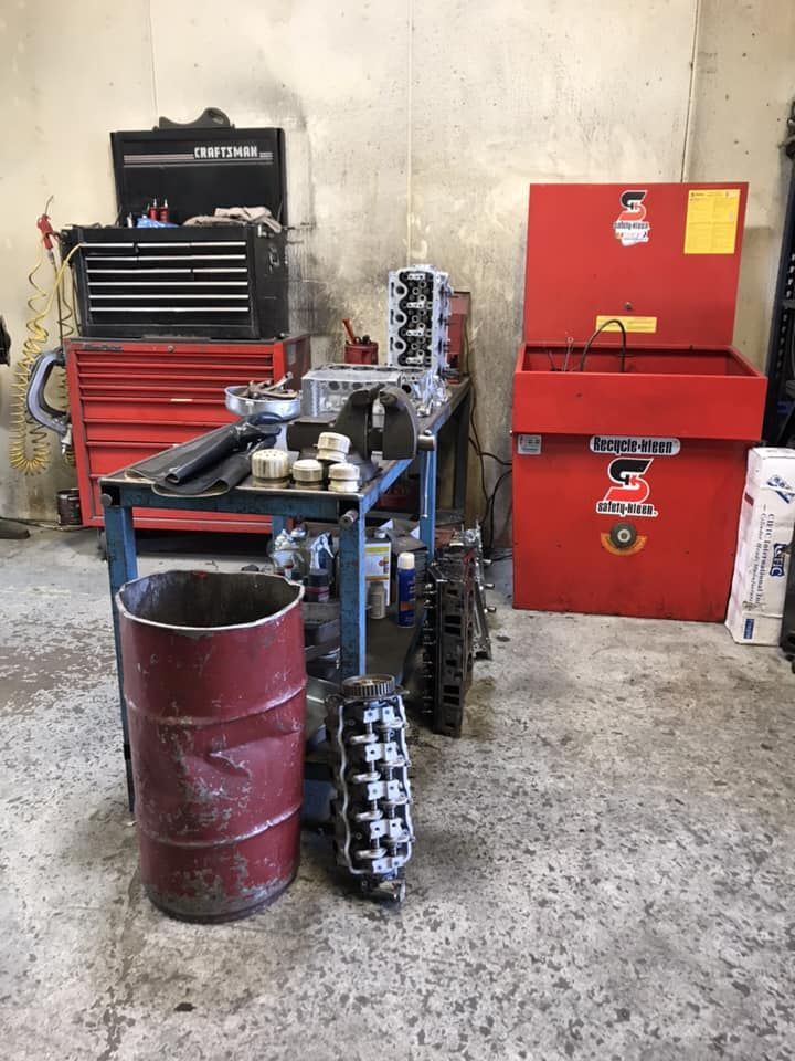 A cluttered automotive repair shop with a workbench, engine parts, a red tool cabinet, and a red parts-washing station.