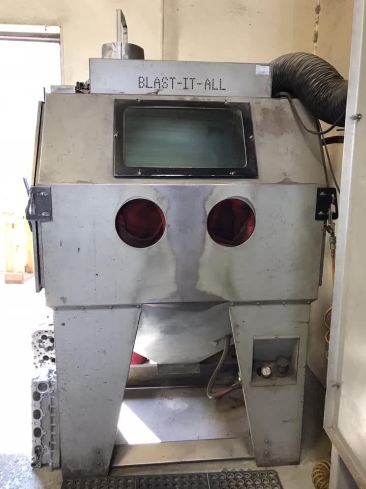 A Blast-It-All brand industrial sandblasting cabinet with a glass viewing window and two glove ports, set in a workshop.