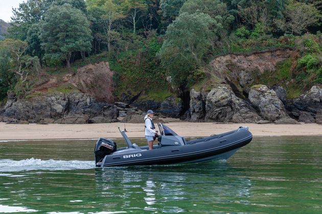 Person steering a small inflatable boat on calm water near a rocky, tree-lined shore