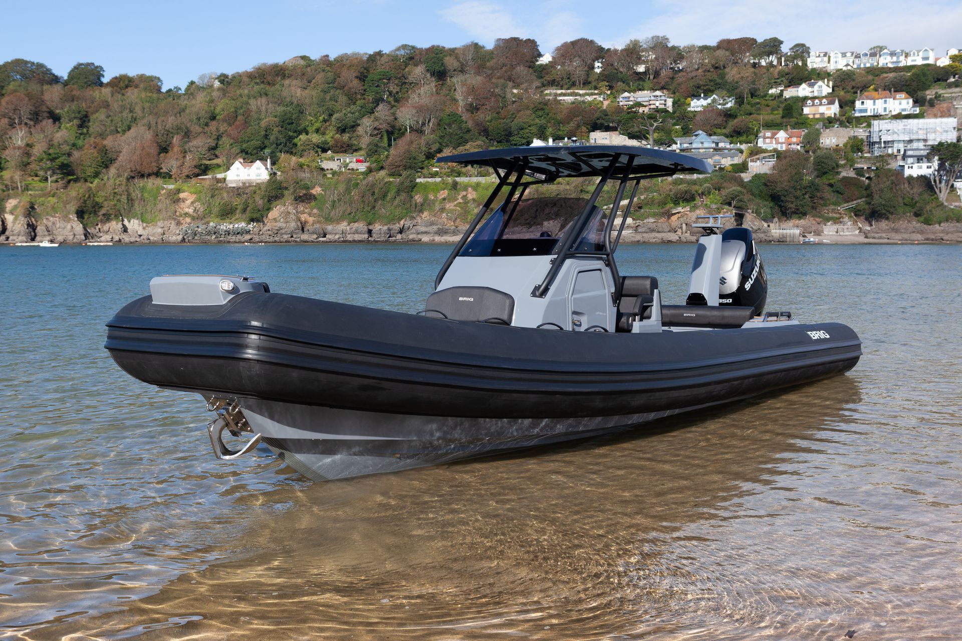 Black inflatable speedboat on a shallow beach by calm water, with wooded hills and houses in the background