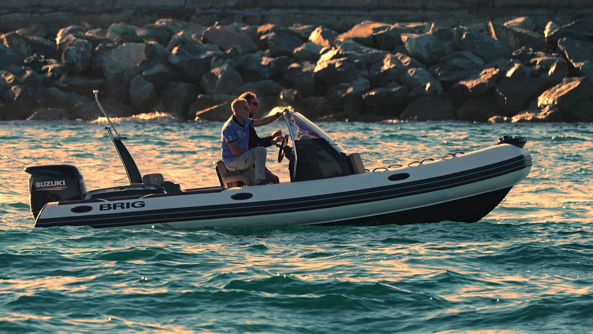 Two people in a small motorboat on choppy water near a rocky shoreline at sunset.