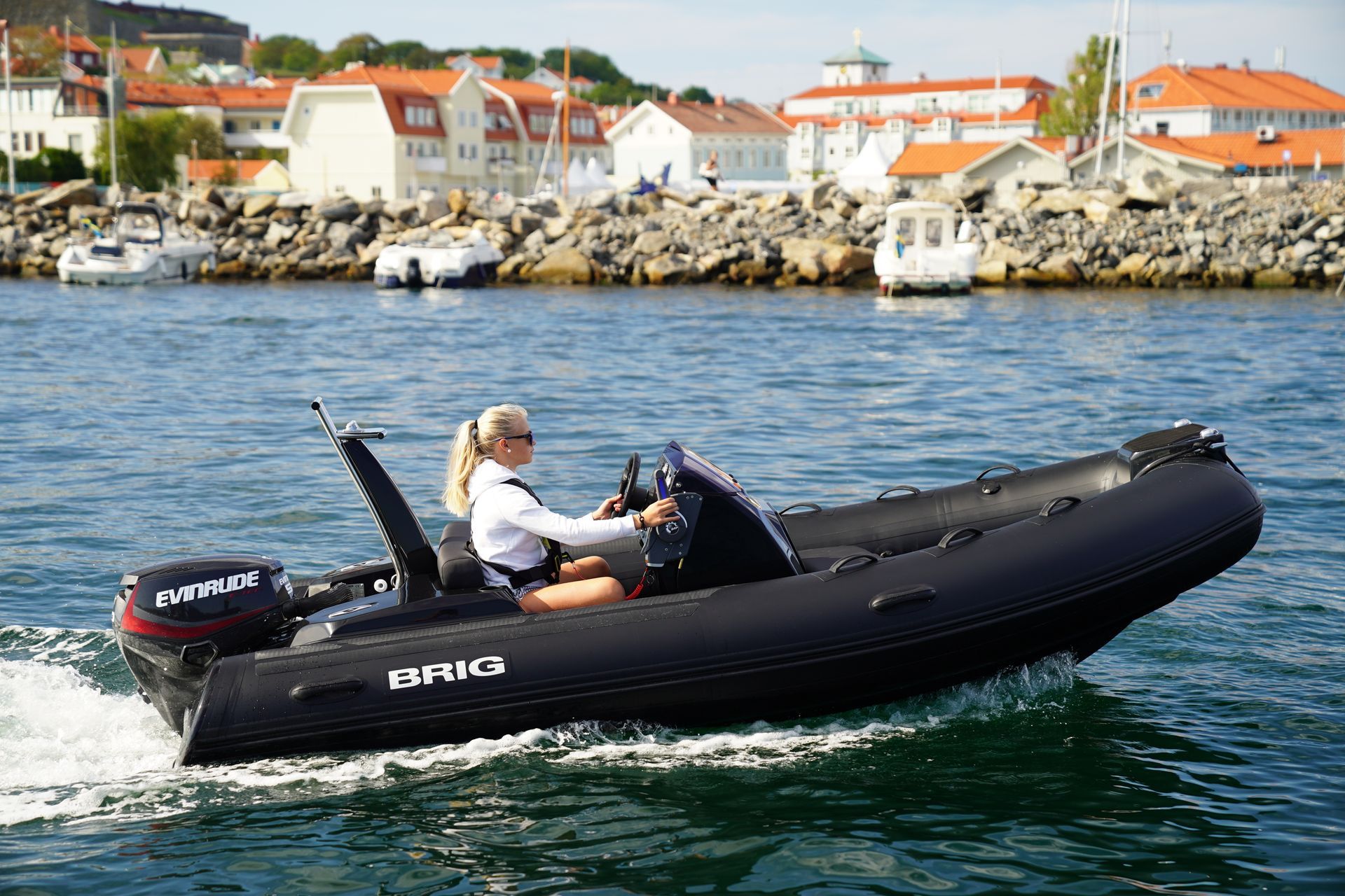 Black inflatable speedboat with a driver on calm water near a rocky harbor and red-roofed houses