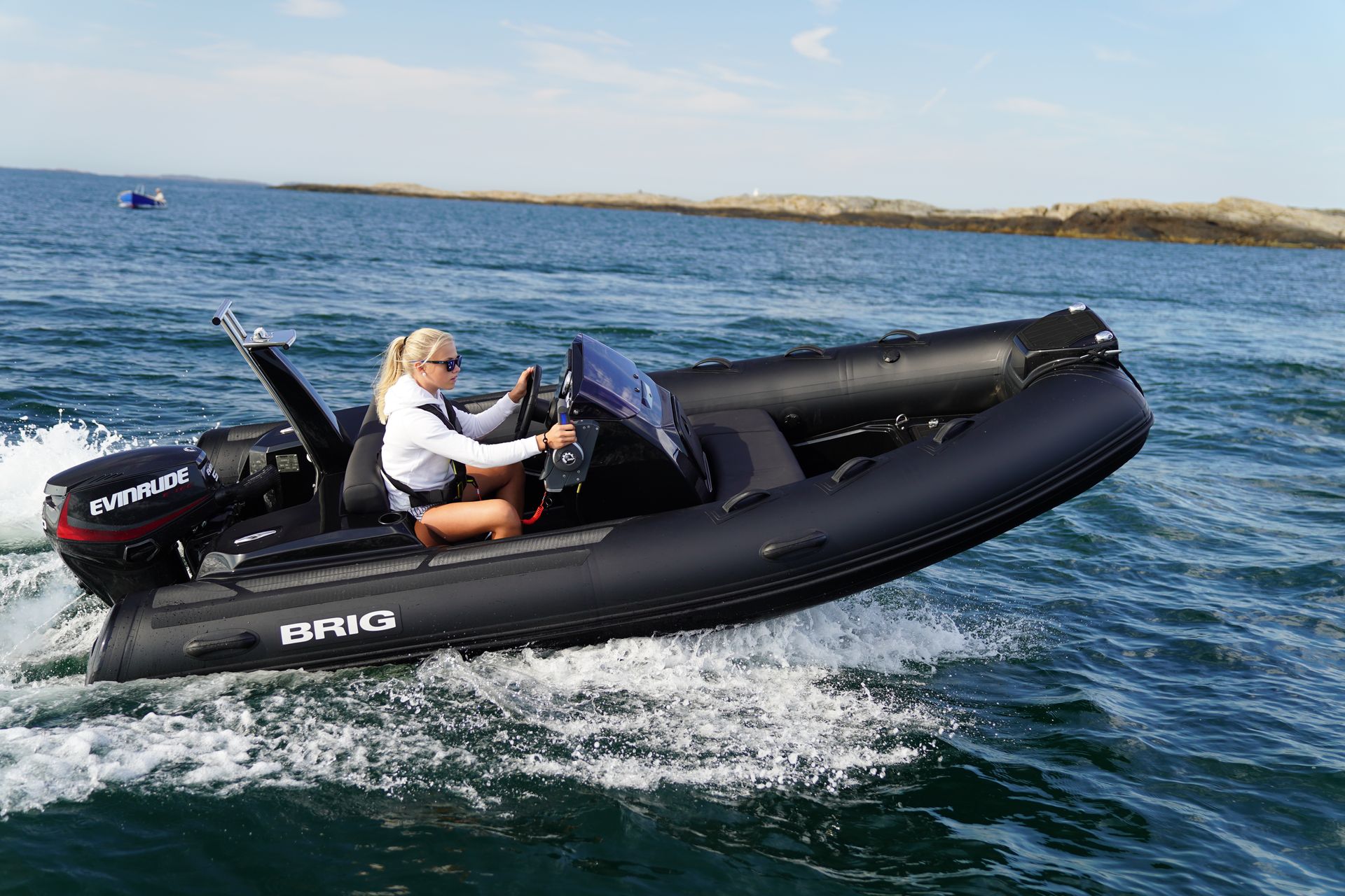 Two people riding a black inflatable motorboat on choppy blue water near a rocky shoreline.