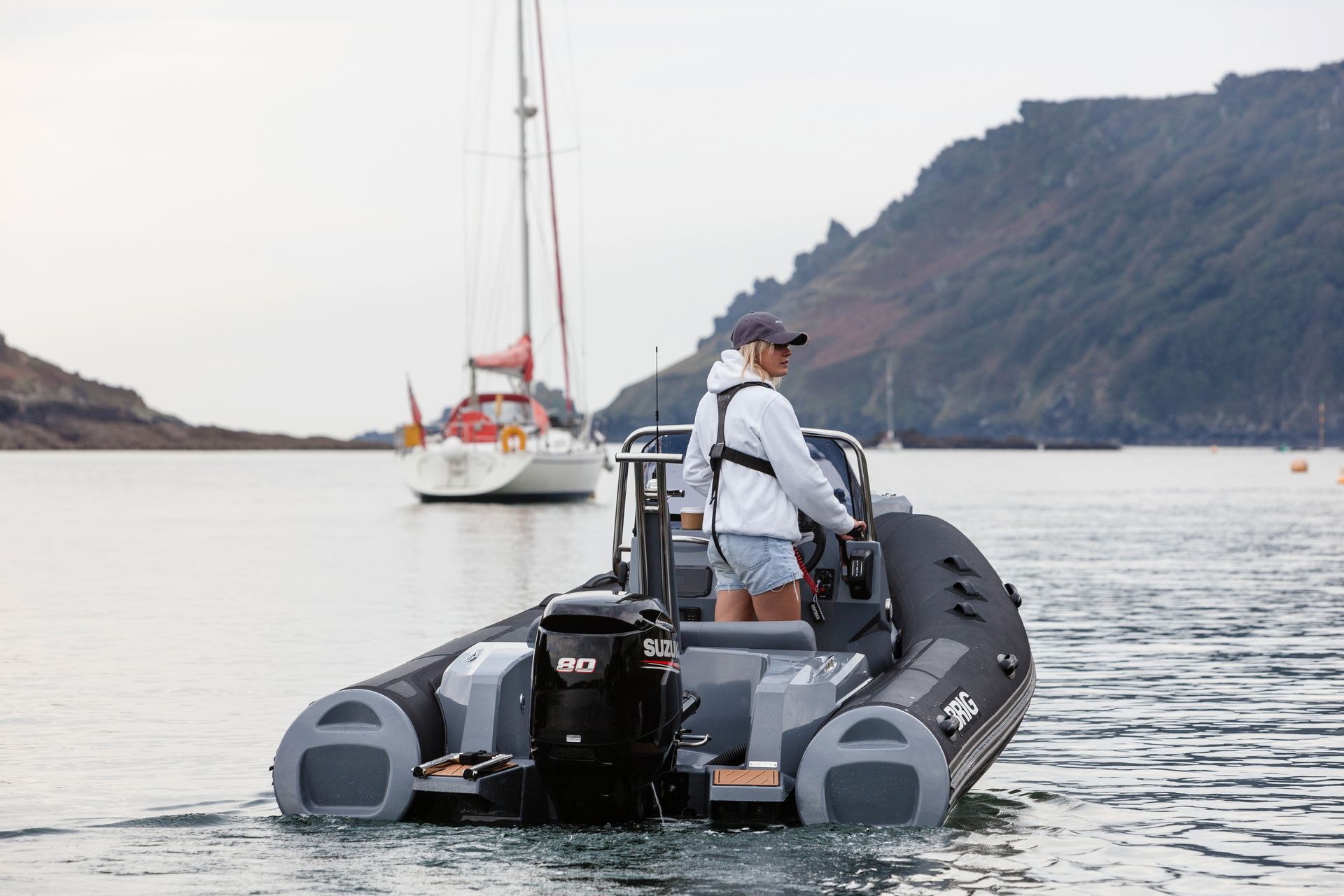 Person in a gray jacket stands on a black inflatable boat on calm water near sailboats and rocky cliffs.