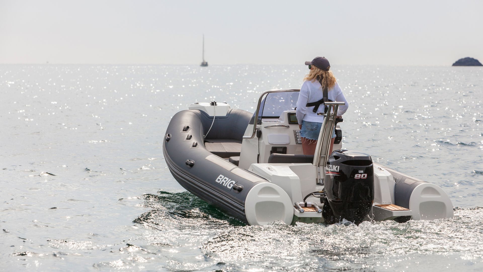 Inflatable motorboat on open water with two people aboard, seen from behind near a small sailboat.