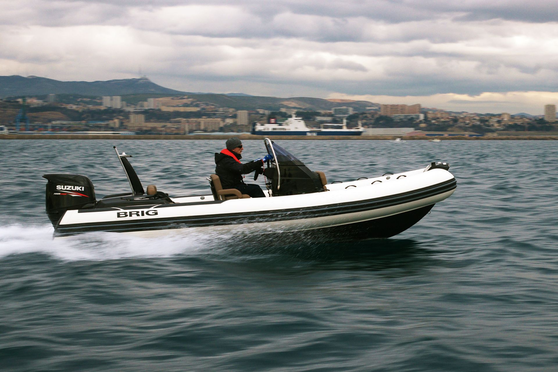 Black-and-white speedboat on calm water with two people, city shoreline and cloudy sky in the background