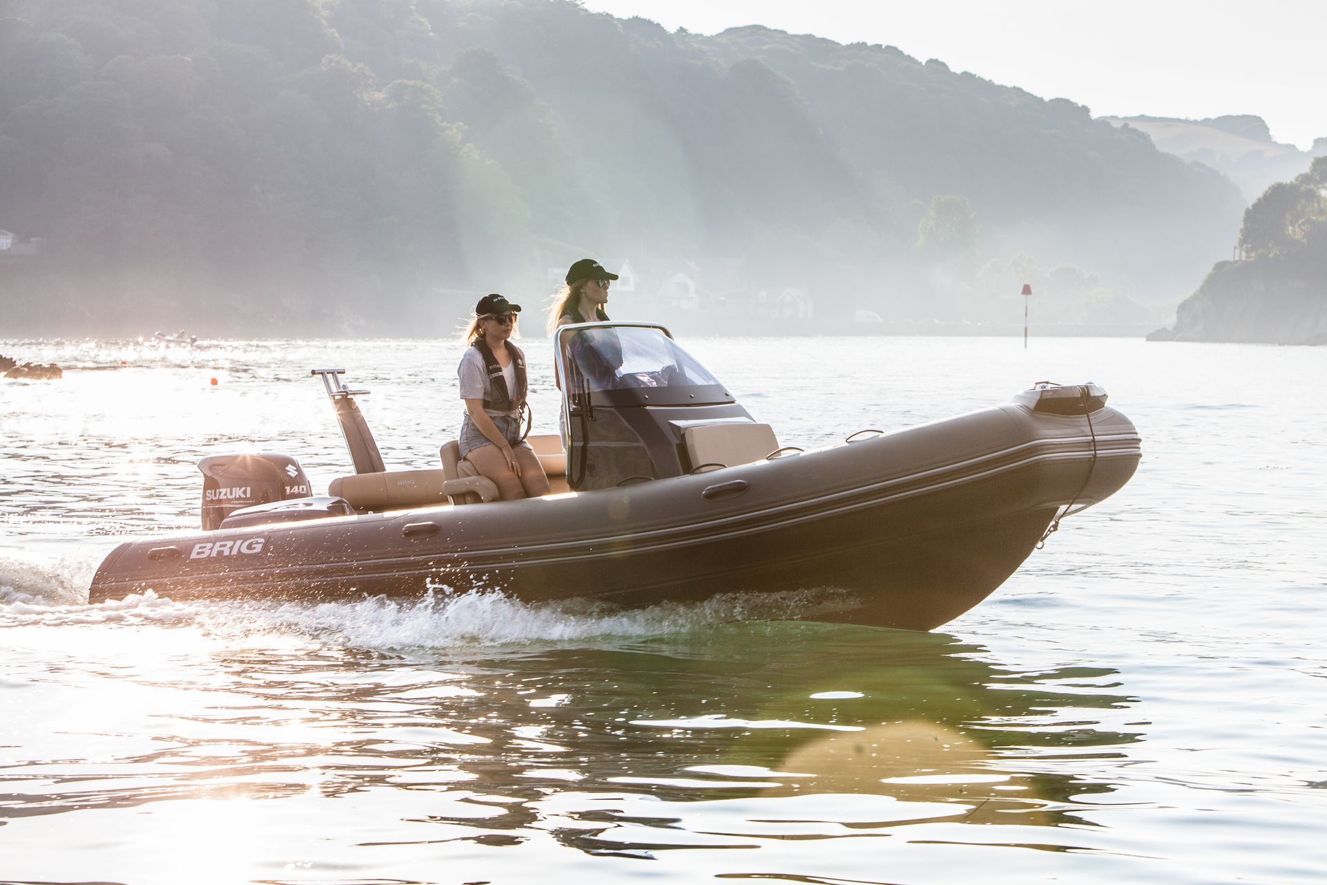 Two people ride a small motorboat on a calm lake at sunrise, with misty hills in the background.