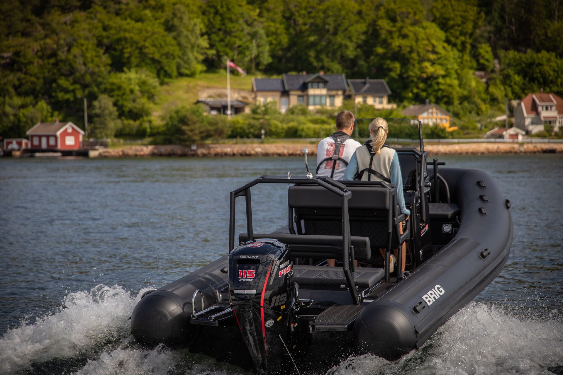 Inflatable black motorboat speeding on a lake with two people aboard, wooded shoreline in background