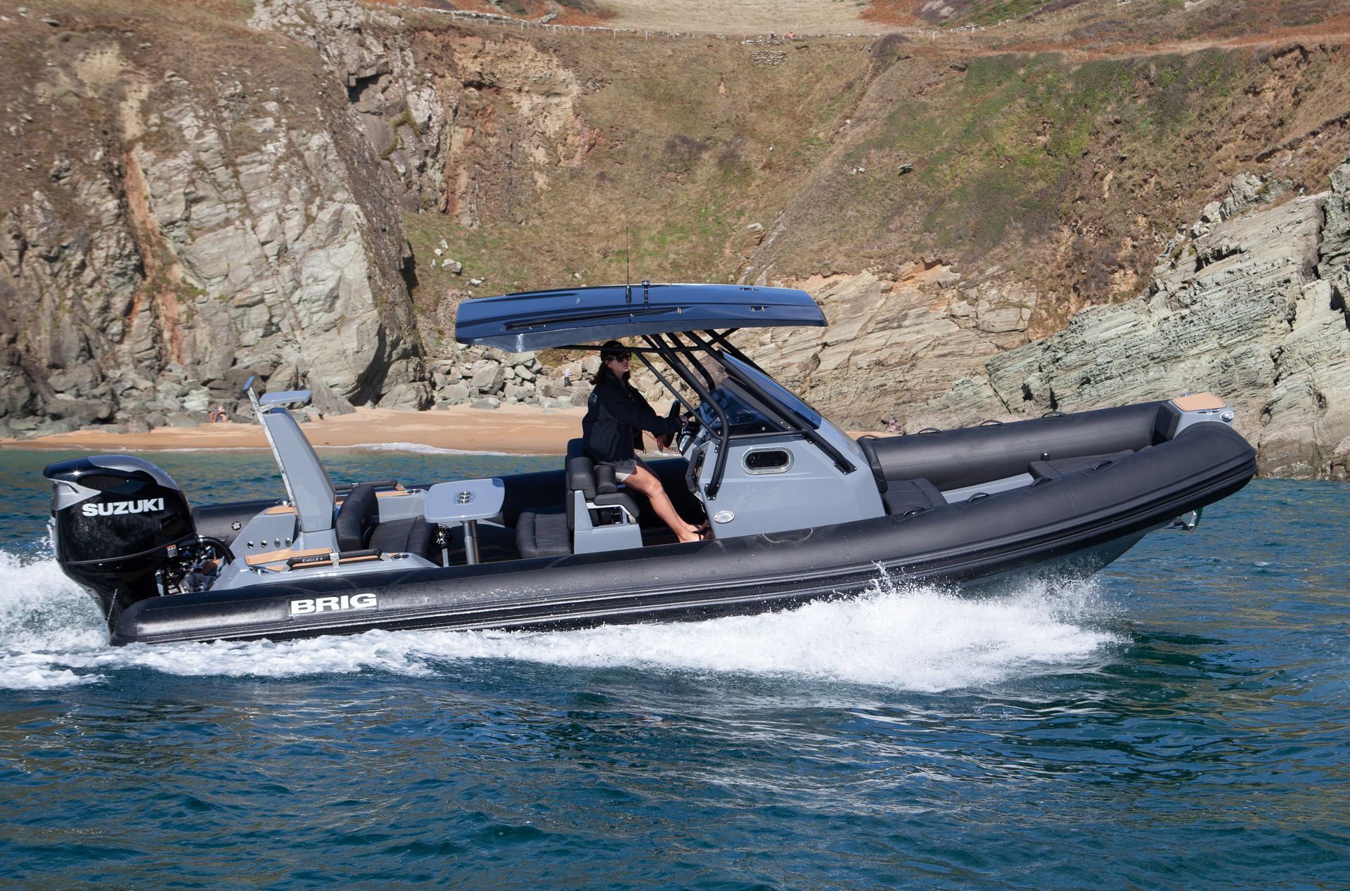 Black inflatable motorboat speeding on blue water near rocky shore, with a blue canopy and seated passenger