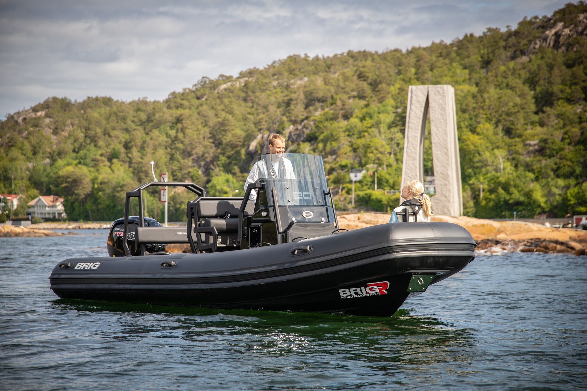 Black inflatable boat with two people on calm water near a rocky shore and wooded hillside.