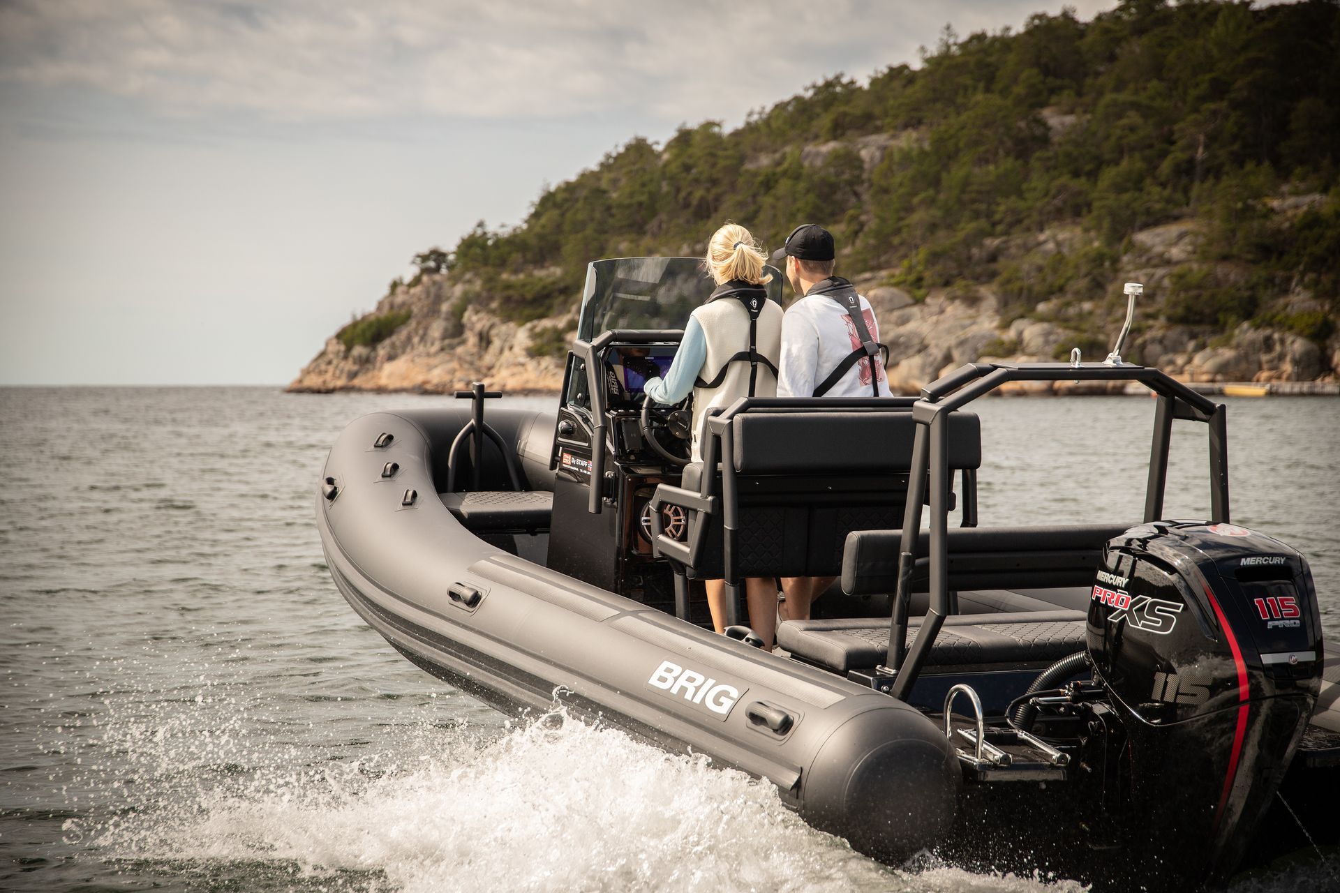 Two people ride a gray inflatable boat on choppy water near a rocky, tree-covered shoreline.