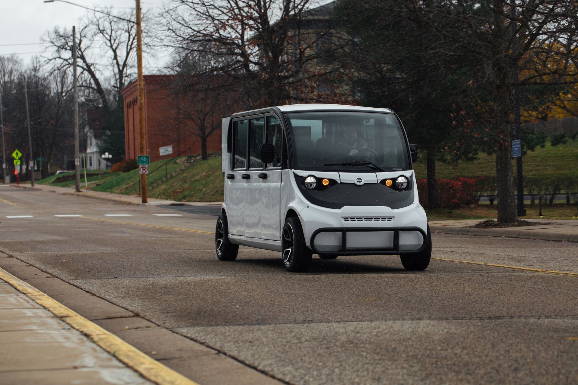 Small white self-driving shuttle driving on a wet street near a brick building and trees