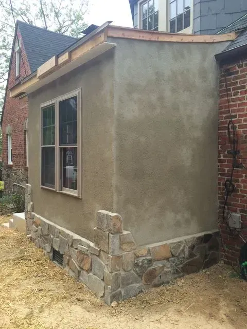 Exterior view of a home addition under construction, with stone foundation, stucco walls, and a window.