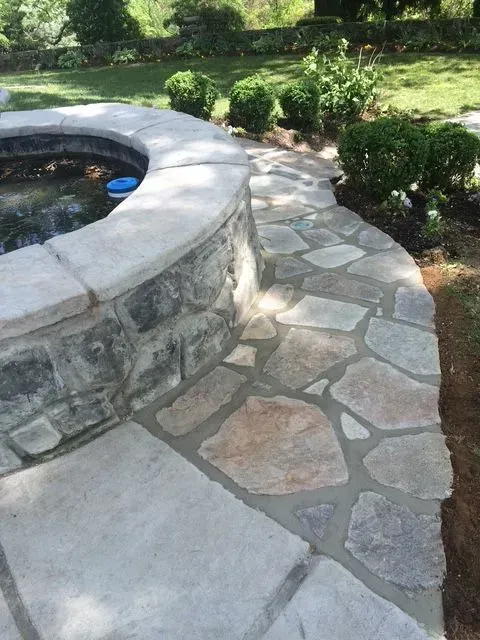 Stone pathway next to a curved stone structure with water feature. Landscaped garden surrounds.