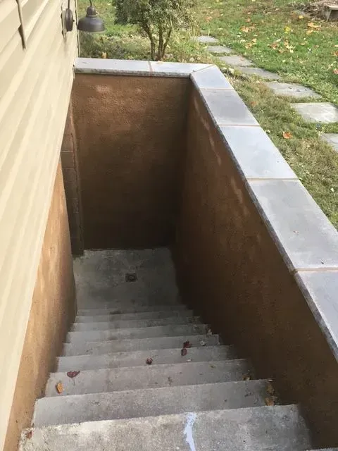 Concrete steps descend into a basement entry with gray stone coping. Tan stucco walls.