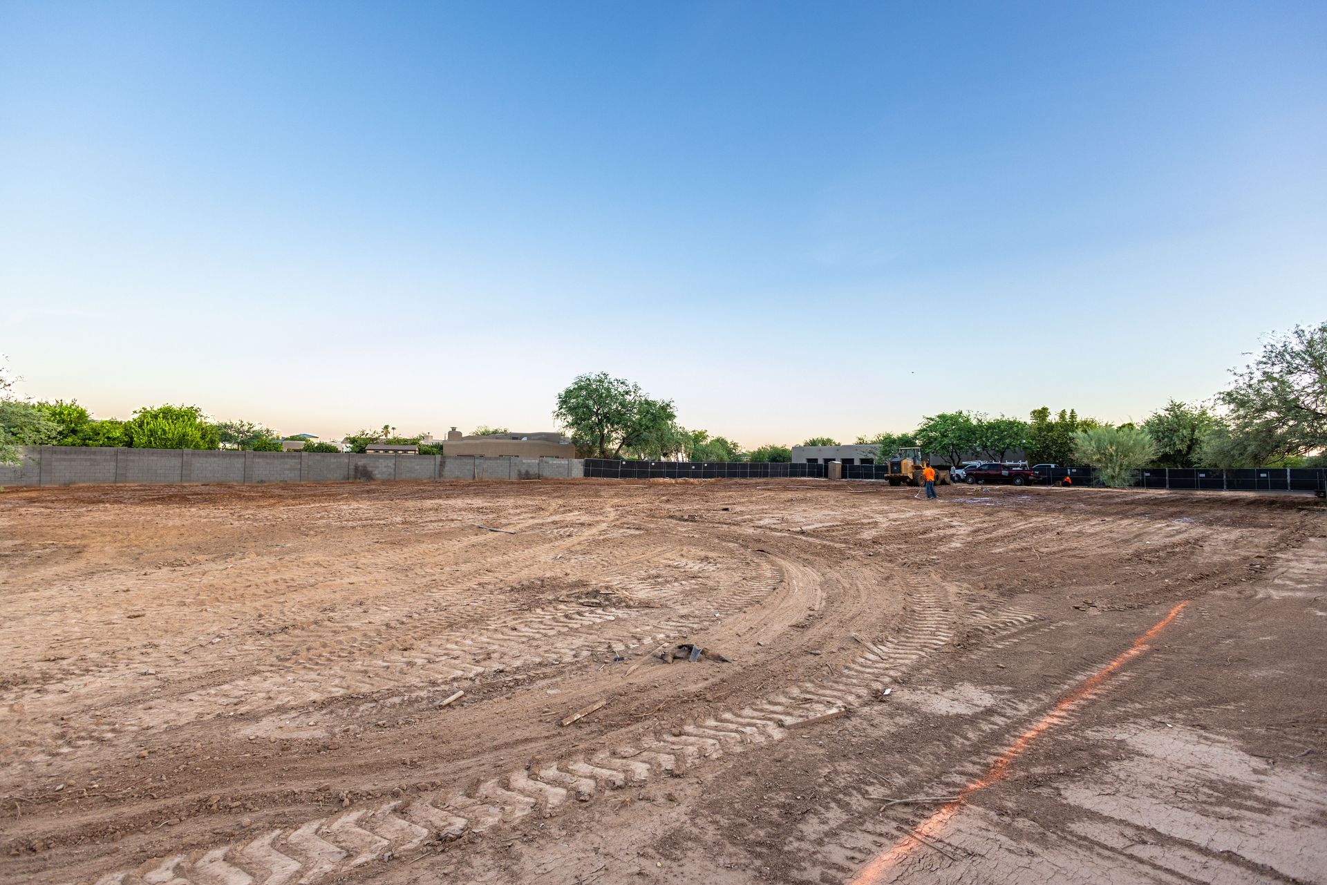 Wide dirt lot with tire tracks, a fence, trees, and a small excavator under a clear blue sky.