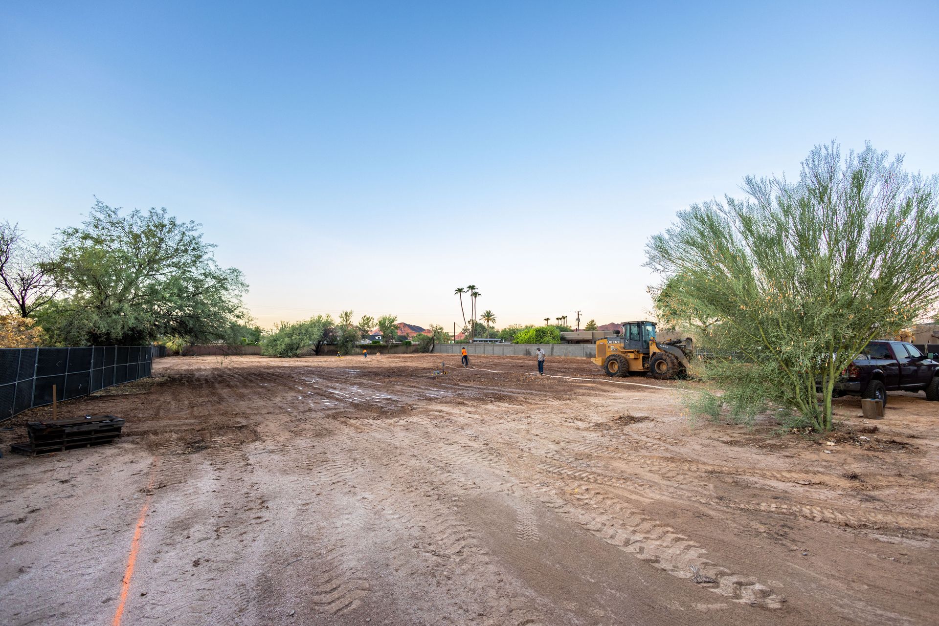 Wide dirt lot at sunset with trees and parked vehicles in the background