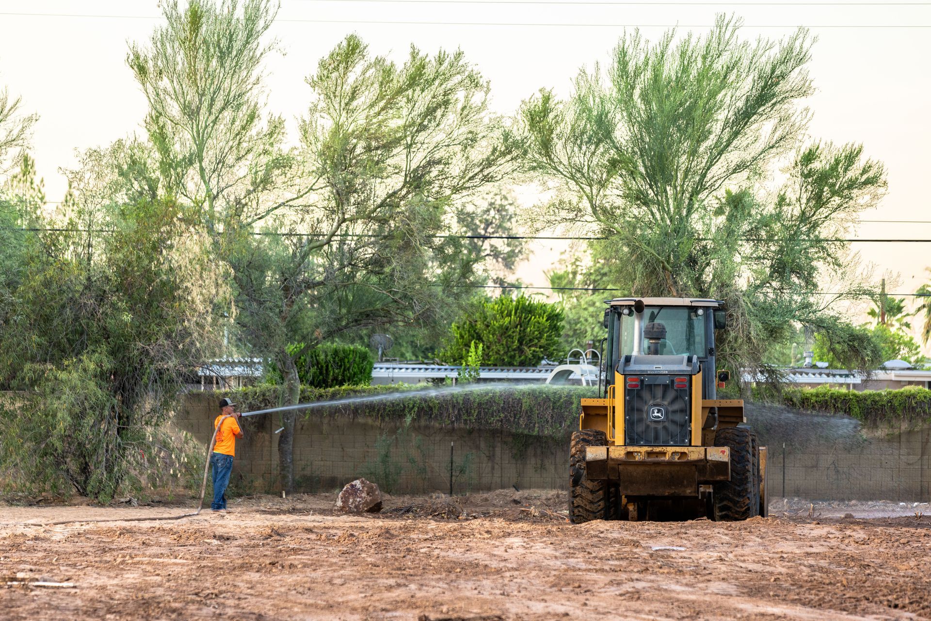 Bulldozer grading muddy ground beside a worker in orange, with trees and a water channel behind.