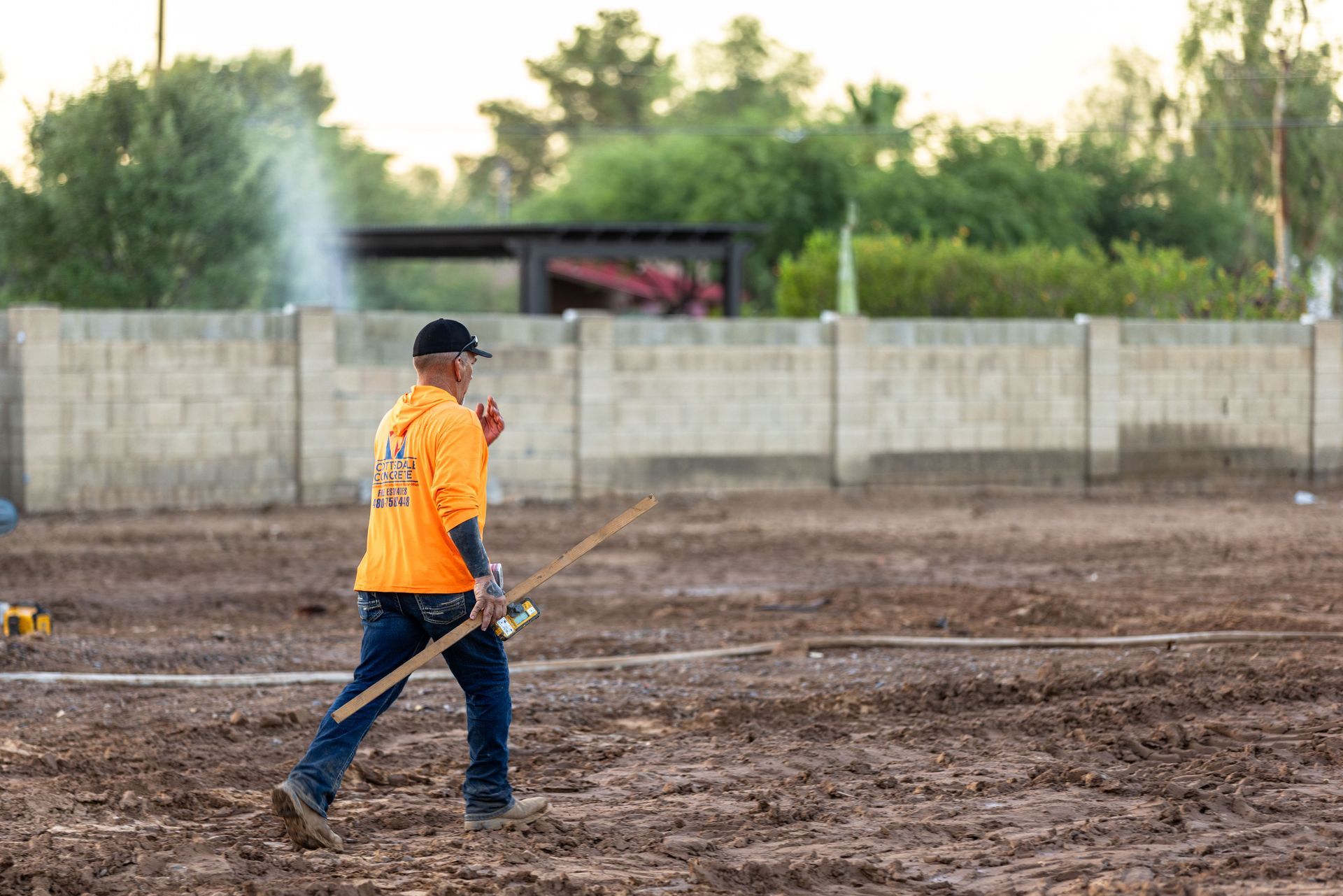 Worker in orange shirt spraying a muddy field near a concrete wall and trees