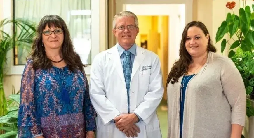 Three people in a hallway. A doctor in a lab coat stands between two women, all smiling.