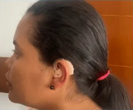 Woman with hearing aid, dark hair pulled back, wearing a black earring, set against a blurred background.