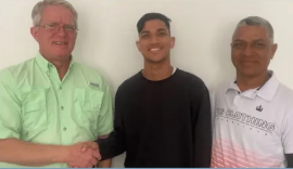 Three men pose against a white wall; one shakes the hand of another, all smiling.