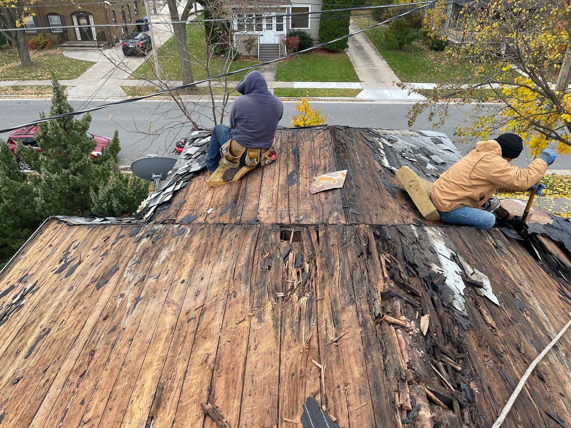 Two workers removing old roofing on a residential roof, overlooking a street with houses and autumn trees.
