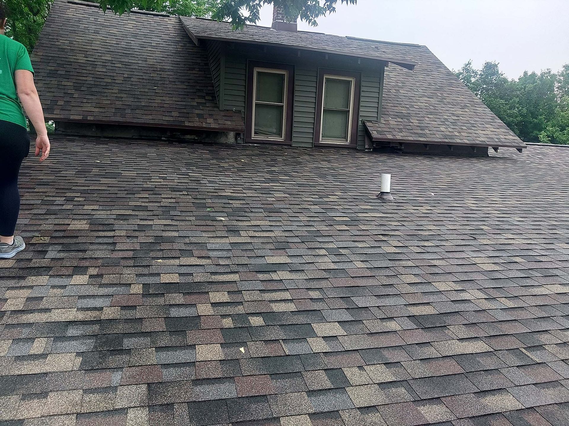 Person on a shingled roof, near a dormer with two windows. Gray and brown shingles. Overcast sky.