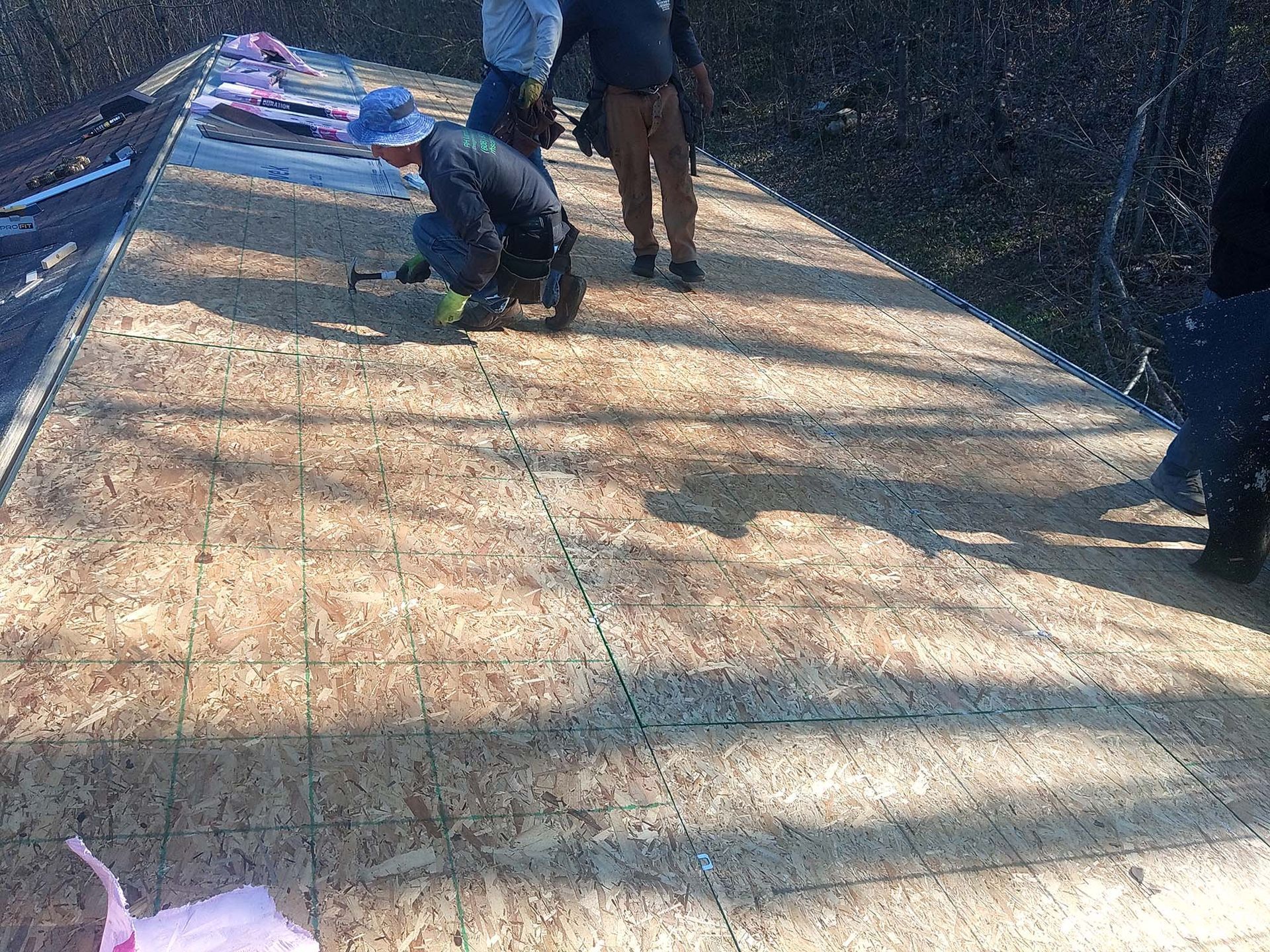 Workers installing roofing on a wood-sheathed roof.  One kneels, using a power tool, while others stand nearby.