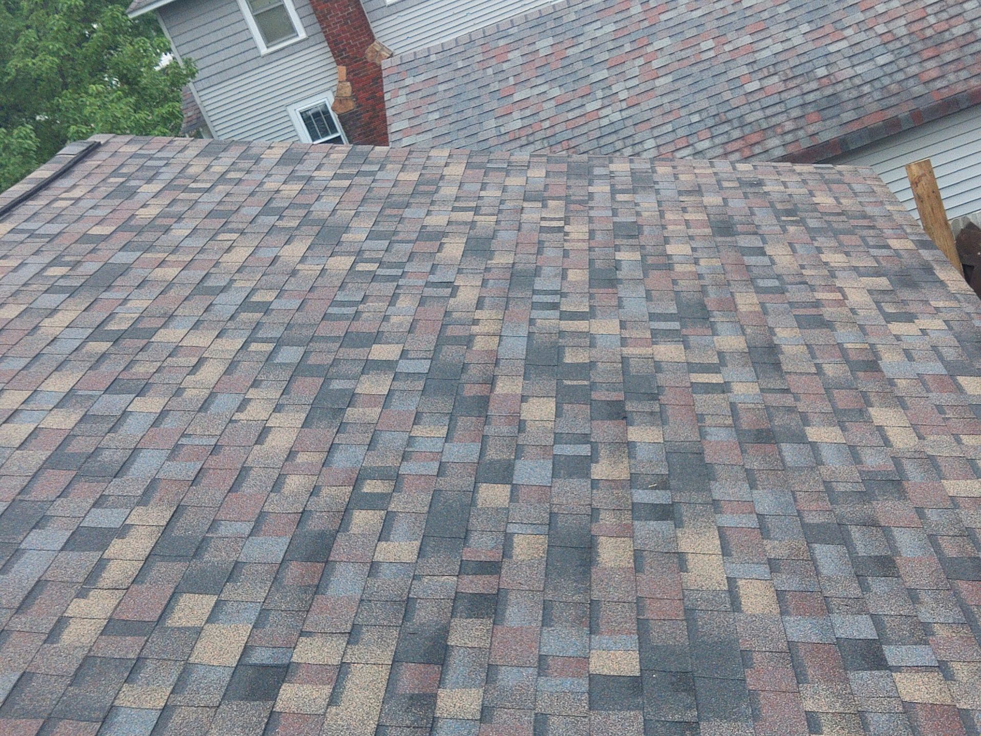 Overhead view of a roof with brown and gray asphalt shingles. A portion of the house is visible in the background.