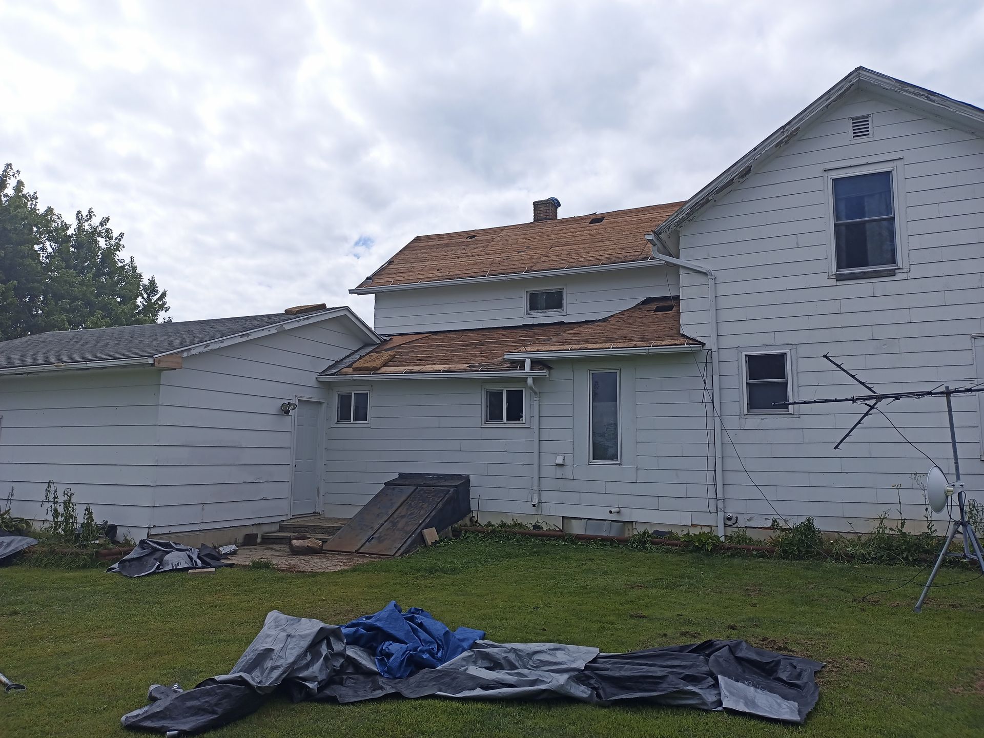 Back view of a white house with damaged roofing and a weathered, sloping ramp in the yard, under a cloudy sky.