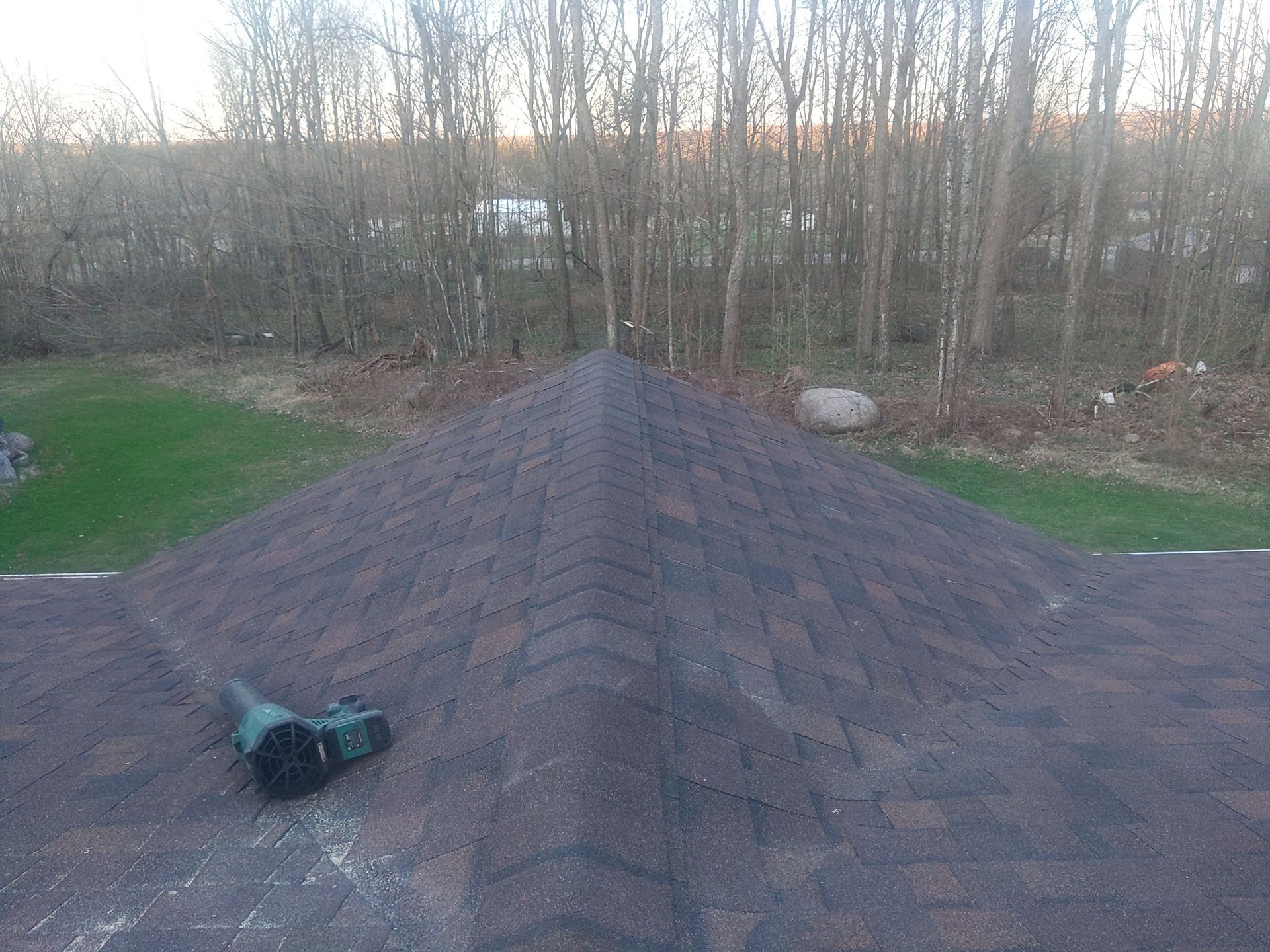 Roof with brown shingles and a green tool, set against a backdrop of trees and grass.
