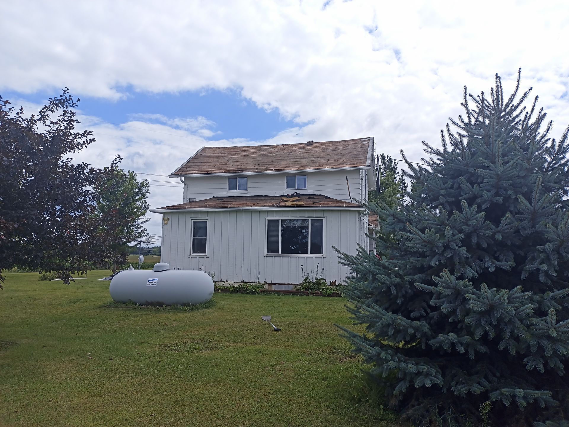 White two-story house with damaged roof, propane tank, blue spruce, and overcast sky.