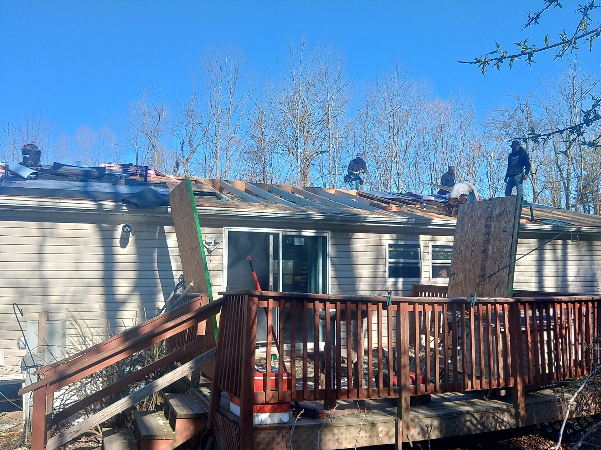 Workers on a roof removing shingles, with a deck in front of a house, trees in background.