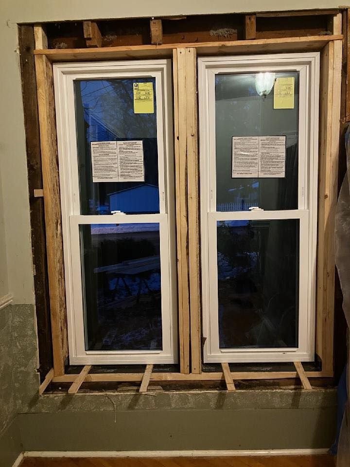 Two white double-hung windows installed in a rough wooden frame within a room's wall opening.